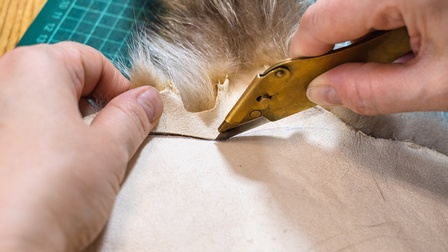 Nahaufnahme zweier Hände einer Person, unter denen ein helles Stück Stoff ist. Am Rand des Stoffes sind kurze, dichte Haare in den Farben weiß, hellbraun und dunkelbraun. Die rechte Hand der Person hält ein spitzes Messer auf die helle Seite des Stoffs