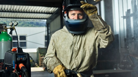 Ein Mann in Schutzausrüstung mit Schweißgerät hebt das Visier seines Helms und lächelt direkt in die Kamera