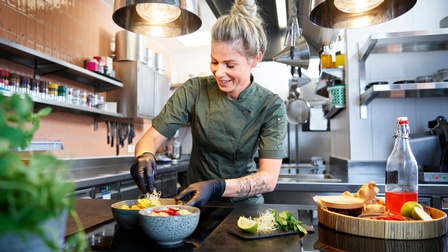 Person mit blonden geschlossenen Haaren und dunkelgrüner Arbeitsbekleidung steht in einer Gastronomieküche und bereitet Speisen zu