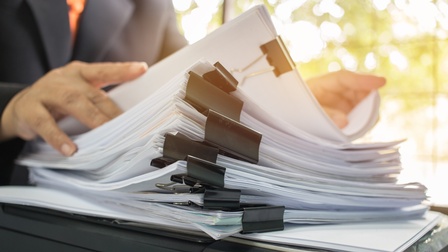 Vor einer Person steht auf einem Tisch ein Papierstapel. Die Person blättert durch den Stapel