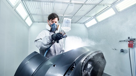 Ein Karosseriebautechniker sprüht ein Autoteil mit einer Flüssigkeit ein. Er trägt eine Atemschutzmaske.