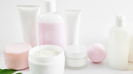 Verschiedene Verpackungen für Kosmetika wie Tuben, Tiegel oder Fläschchen aus Plastik ohne Etikett, ein Tiegel mit Puder ist geöffnet, eine rosa Badekugel sowie Blätter liegen daneben