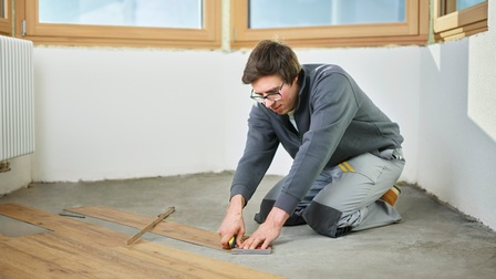 Person in grauer Arbeitsmontur kniet auf Boden und steckt Holbodenbretter zusammen, im Hintergrund Heizkörper und Fenster