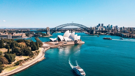 Ansicht einer Stadt im Sonnenschein unter blauem Himmel: Mittig modernes Gebäude mit segelartigen Dächern an großem Stadtgewässer liegend, auf dem Boote verkehren, im Hintergrund große Brücke mit Stahlkonstruktion und Gebäude