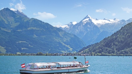 Ein Boot mit einem Glasdach sowie zwei Österreichflaggen vorne und hinten fährt über einen See. Im Hintergrund sind teilweise schneebedeckte Berge sowie der blaue Himmel mit vereinzelten Wolken
