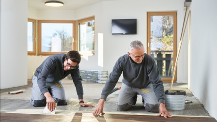 Zwei Personen in grauen Arbeitsmonturen knien am Boden und verlegen Holzbodenbretter, im Hintergrund Fenster, Balkontüre und Fernseher