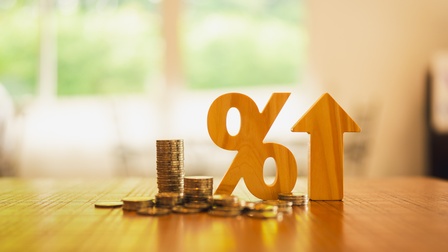 Auf einem Tisch stehen bzw liegen direkt nebeneinander: Münzstapel, verstreute Münzen, ein hölzernes Prozentzeichen, ein hölzerner Pfeil nach oben