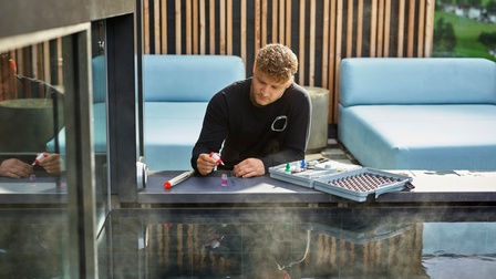 Person mit kurzen braunen Haaren und Bart trägt einen schwarzen Pullover und begutachtet eine Flüssigkeit bei einem beheizten Pool, daneben liegt ein Koffer mit weiteren Behältern sowie ein Thermometer am Beckenrand, dahinter ein Entspannungsbereich