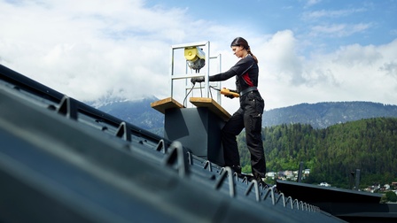 Eine Frau arbeitet an einem Kamin. Im Vordergrund das schwarze Schrägdach. Im Hintergrund eine Bergkulisse