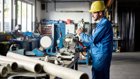 Ein Mann in der Werkstatt schneidet dicke Plastikrohre. Er trägt Blaumann und einen gelben Helm.