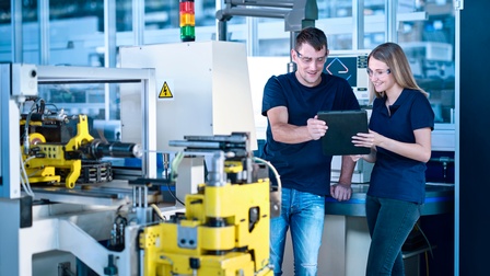 Zwei lächelnde Personen mit Schutzbrillen halten gemeinsam großes Tablet und blicken darauf, ringsum industrielle Geräte