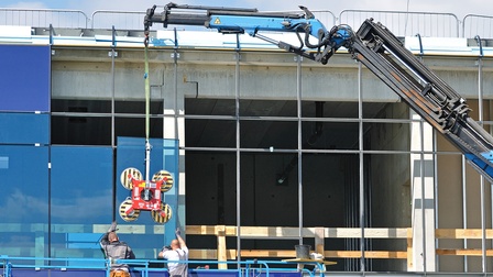 Zwei Personen auf einer Plattform vor einer teilweise verglasten Fassade. Über ihnen ist der Arm eines Krans. Daran hängt eine Hebevorrichtung mit Saugnäpfen, die an einer Glasscheibe befestigt ist. Die Personen führen die Scheibe in Richtung der Fassade
