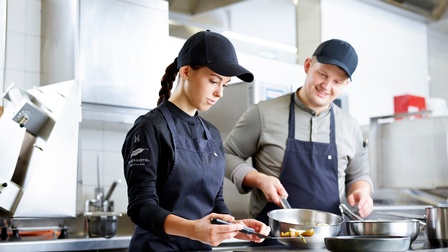 Zwei Personen mit Caps und in Schürzen stehen in Edelstahlküche: Eine Person schwenkt Pfanne, die andere hievt mit Hausfreund gegrilltes Gemüse von Platte