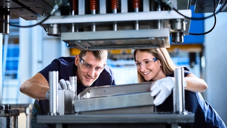 Zwei lächelnde Personen mit Schutzbrillen und Handschuhen beugen sich über industrielle Maschine