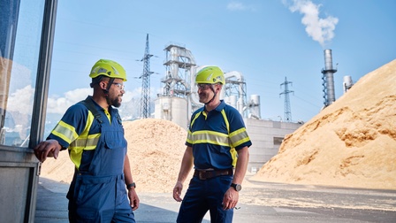 Zwei Personen mit gelben Schutzhelmen, Schutzbrillen und in blau-gelber Arbeitsmontur stehen auf Platz einer Industrieanlage, im Hintergrund Strommasten, industrielles Gebäude und zwei große Haufen gehäckseltes Holz 