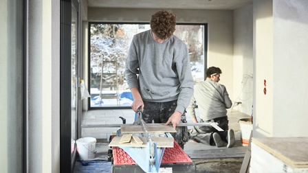 Ein junger Mann schneidet Fliesen - im Hintergrund werden sie von seinem Kollegen verlegt. 
