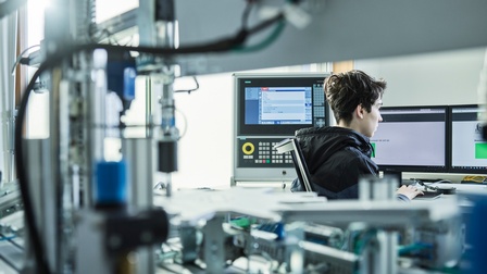 Ein junger Mechatroniker arbeitet am Computer. Im Vordergrund Elektronik und Maschinen