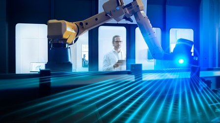 Person mit Brillen und in weißem Laborkittel blickt durch Glasscheibe auf Roboterarm mit blauem Laserlicht