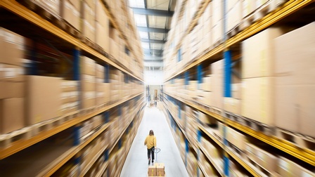 Lagerhalle mit hohen Regalen in Bewegungsunschärfe, in der Mitte Person die Palettenhubwagen mit darauf platzierten Schachteln hinter sich herzieht