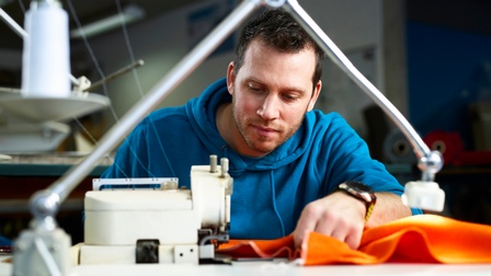 Ein junger Mann sitzt an der Nähmaschine und vernäht orangenen Stoff