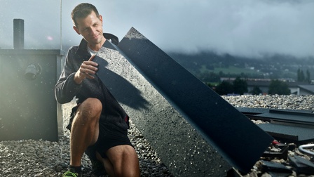 Ein Spengler auf einem Flachdach betrachtet ein Blechteil zur weiteren Verarbeitung. Im Hintergrund eine nebelige Landschaft