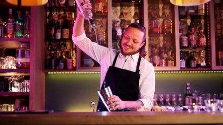 Person mit  schulterlangen nach hinten gefassten Haaren, Bart, weißem Hemd und dunkler Schürze steht hinter einer Theke vor einem Spirituosenregal und gießt eine Flüssigkeit von einer Spirituosenflasche in einen Cocktailshaker