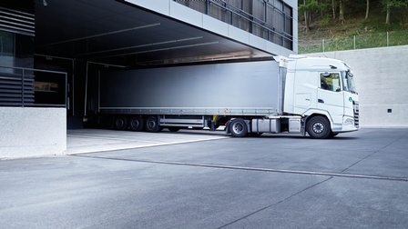 Weißer LKW steht bei einem Lagerkomplex, im Hintergrund zeigt sich ein angrenzender Wald oberhalb einer Mauer