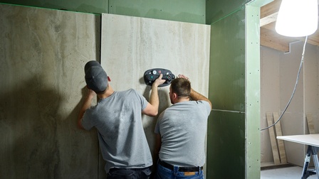 Zwei Männer fixieren eine sehr große, graue Fliese an einer Rigips-Wand.