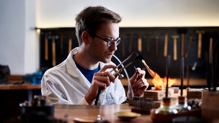 Ein junger Mann in der Werkstatt lötet etwas auf einen Goldring auf. Mit einem Mundstück kontrolliert er die Sauerstoffzufuhr