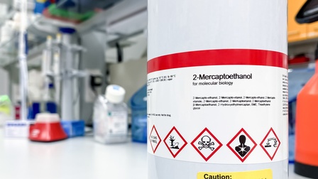 Weißer Metalleimer mit rotweißem Etikett mit schwarzer Aufschrift 2-Mercaptoethanol und verschiedenen weißen Warnzeichen mit roter Umrandung wie Totenkopf, darunter weiteres gelbes Etikett mit Aufschrift Vorsicht, Überdruck möglich