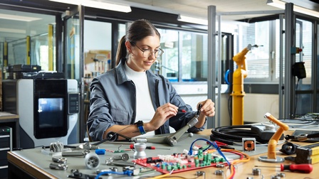 Eine junge Frau schraubt an einer Art Hebel oder Halterung. Im Vordergrund elektronische Teile und Werkzeug