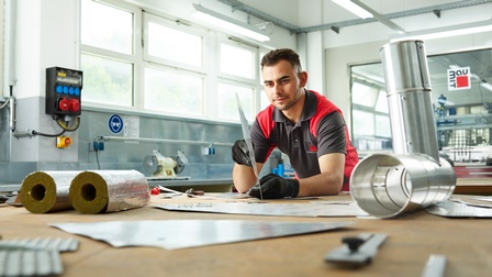 Person mit schwarzen Arbeitshandschuhen steht in Werkstatt und inspiziert Metallplatte über Werktisch gebeugt