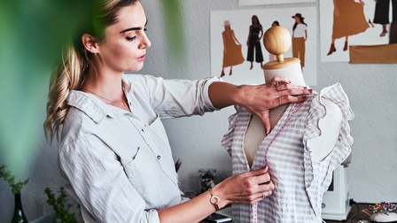 Eine Person mit hellen Haaren steht seitlich neben einer Modepuppe, auf der ein gestreiftes Oberteil hängt. Die Person hält mit beiden Händen ein Maßband von der linken Schulter der Puppe über die Brust und blickt darauf