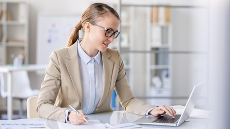 Person in Businesskleidung, geschlossenen Haaren und Brille sitzt an einem Schreibtisch, blickt auf einen Laptopbildschirm und macht sich Notizen