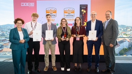 Foto von sieben Personen, die nebeneinander vor einer Präsentationsfläche stehen. Vier Personen in der Mitte halten Auszeichnungen: Zwei halten Urkunden, zwei weitere jeweils eine Trophäe mit Sternmotiv. Eine weitere Person hält ebenfalls eine Urkunde. Auf der Präsentationsfläche steht das Logo „WKO – DER HANDEL“