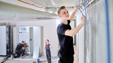 Ein junger Mann verlegt Kabel im Rohbau an der Decke. Im Hintergrund eine Kollegin und ein Kollege am Schaltkasten