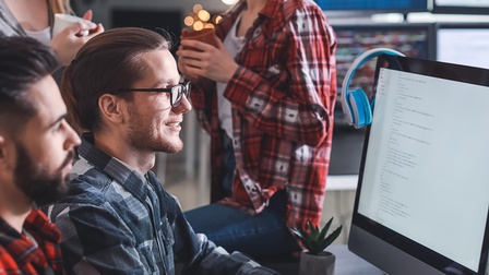 Seitliche Aufnahme zweier neben einem Bildschirm sitzender Personen. Daneben sind zwei weitere stehende Personen. Alle blicken auf den Bildschirm, auf dem mehrere Zeilen sind