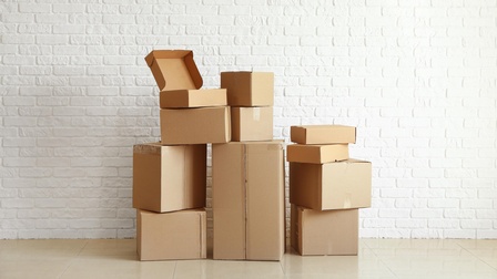 Kartons unterschiedlicher Größe stehen nebeneinander und übereinander gestapelt vor einer weißen Wand auf hellen Fliesen