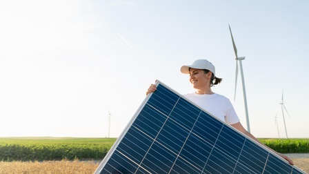 Eine Person trägt ein Solarpanel über ein Feld, im Hintergrund stehen Windräder.