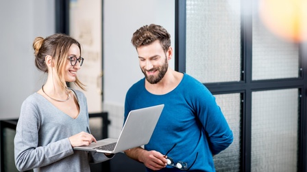 Zwei lächelnde Personen auf aufgeklapptes Notebook blickend