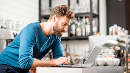 Person mit Bart in blauem Pullover über Laptop gebeugt, im Hintergrund Utensilien einer Bar