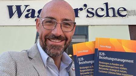 Ein lächelnder Mann mit Brille hält mehrere Exemplare eines Buches in den Händen und steht vor einer Buchhandlung.