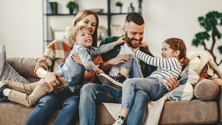Zwei Erwachsene und zwei Kinder sitzen auf einem hellbraunen Sofa. Die Kinder liegen auf den Erwachsenen. Alle interagieren miteinander. 
