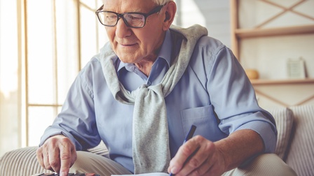 Person mit weißem Haar und Brille bedient mit einer Hand Taschenrechner und notiert mit zweiter Hand in Buch
