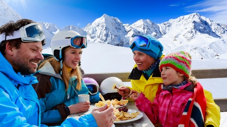 Familie in Wintersportbekleidung speist auf einer Hütte im Freien während sich im Hintergrund ein Bergpanorama bietet 
