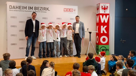 Auf einer Bühne stehen sechs Kinder nebeneinander, alle tragen weiße T-Shirts, rote Weihnachtsmützen und Medaillen um den Hals. Die Kinder lächeln und haben die Arme umeinander gelegt. Links und rechts von der Gruppe stehen zwei erwachsene Männer in Business-Casual-Kleidung.<br />
<br />
Im Hintergrund ist eine große Wand mit dem Schriftzug „DAHEIM IM BEZIRK. Ihre Unternehmerkammer im Bezirk Schwaz.“ sowie mehreren WKO-Logos zu sehen. Rechts auf der Bühne steht ein hoher, farbiger Würfelturm mit den Buchstaben „WKO“ und „WIFI“.<br />
<br />
Vor der Bühne sitzen zahlreiche Kinder auf dem Boden und schauen zur Gruppe hinauf. Einige tragen ebenfalls Weihnachtsmützen oder farbige Pullover. Die Szene wirkt wie eine feierliche Preisverleihung oder Ehrung im Rahmen einer Veranstaltung der Wirtschaftskammer.