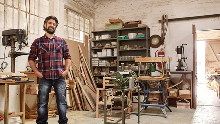 Cooler, lächelnder Tischler mit Bart steht in seiner Werkstatt mit Händen in Hosentaschen