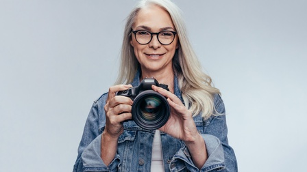 Lächelnde Person mit Brillen hält digitale Kamera in Händen, Hintergrund grau