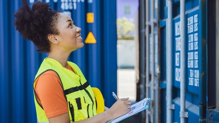 Person mit gelber Warnweste steht fröhlich vor einem Container und notiert sich etwas auf einem Klemmbrett während im Hintergrund weitere blaue Container platziert sind