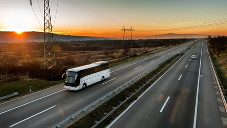 Seitliche Aufnahme eines weißen Autobusses auf einer zweispurigen Autobahn, in deren Mitte eine Abtrennung ist und danach wieder zwei Spuren sind. Im Hintergrund sind braune und grüne Felder sowie Strommasten